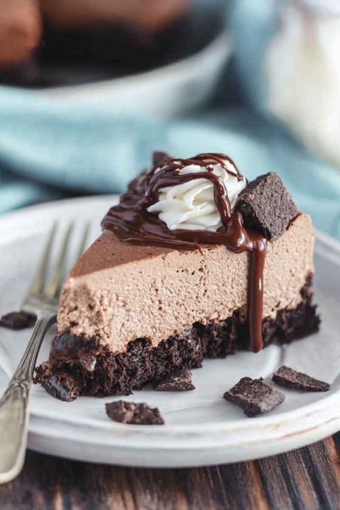 A slice of Brownie Batter Cheesecake with layers of brownie crust, cheesecake filling, whipped cream, crumbled brownies, and hot fudge sauce.