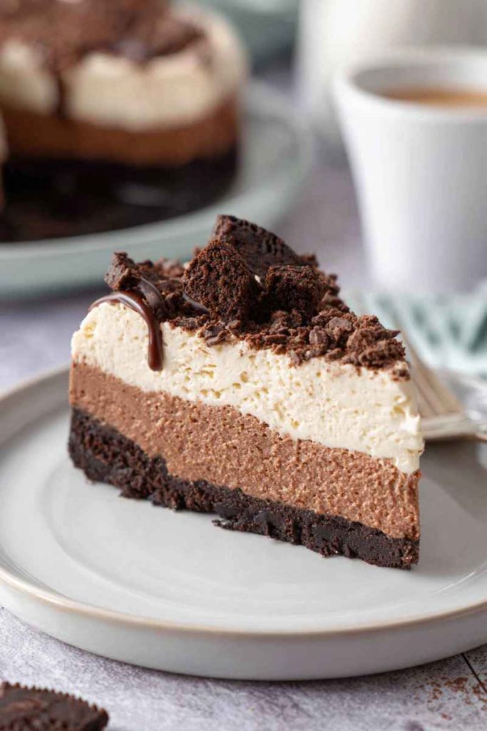 A slice of Brownie Batter Cheesecake with layers of brownie crust, cheesecake filling, whipped cream, crumbled brownies, and hot fudge sauce.
