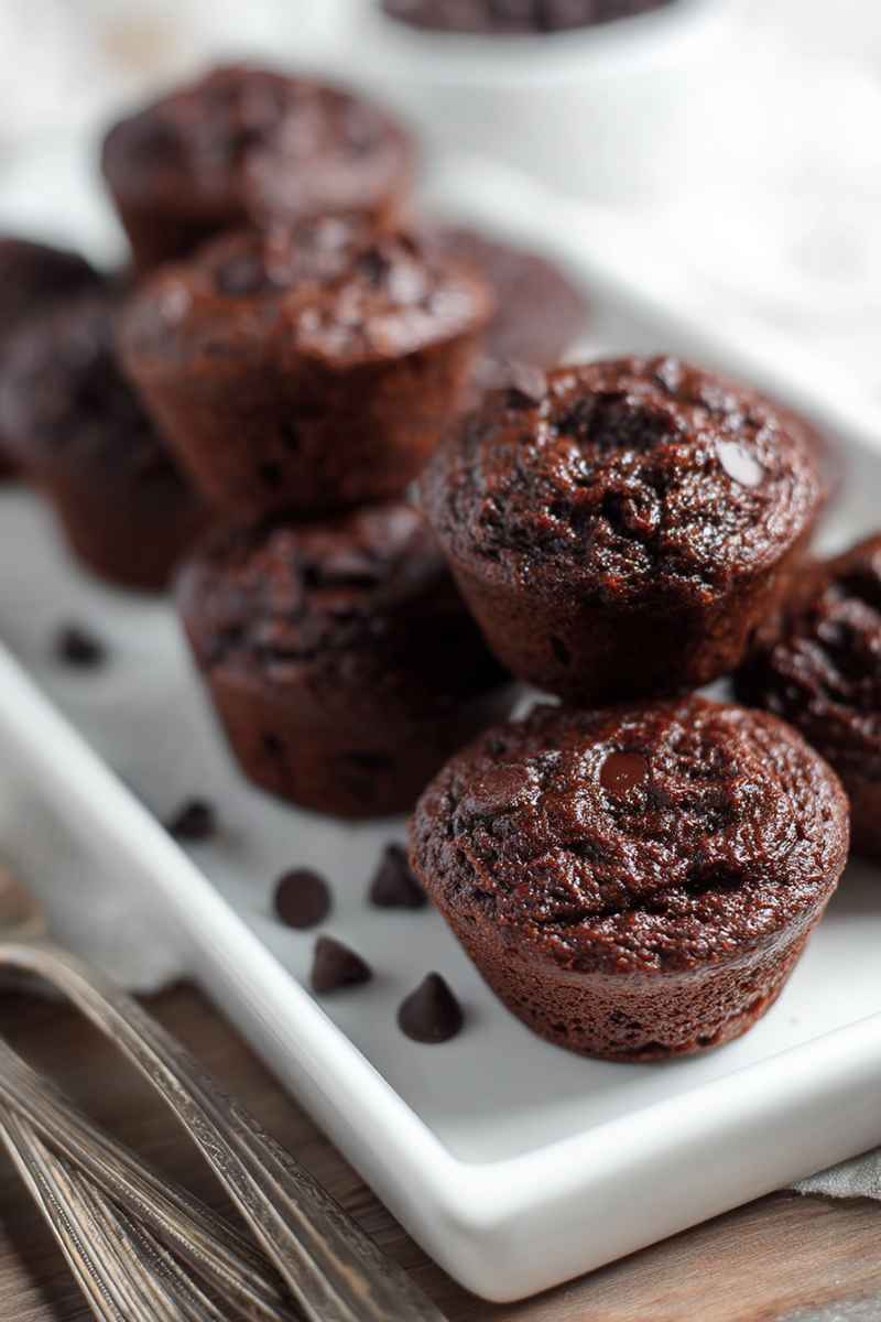 Stacked chocolate muffins with visible chocolate chips, served on a white plate with scattered chips around.