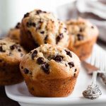 Tall, golden-brown Chocolate Chip Muffins on a white plate, showcasing their moist texture and scattered chocolate chips.