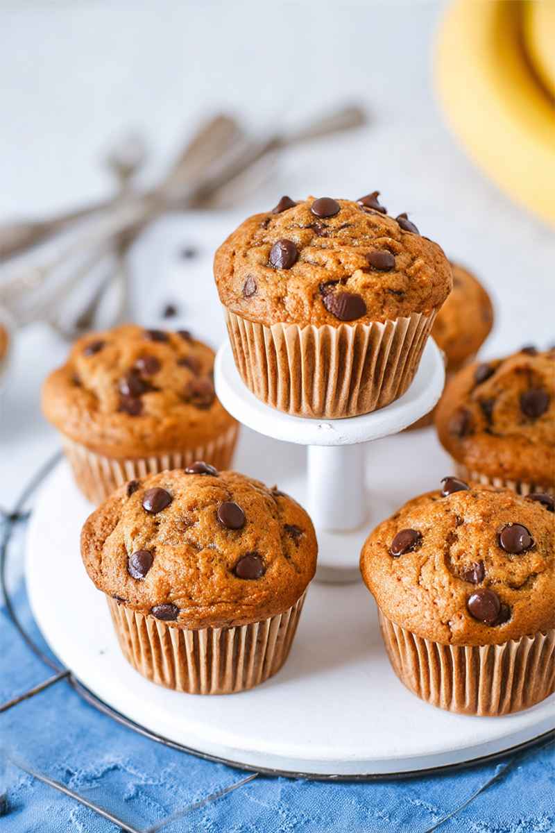 Tiered arrangement of golden-brown banana chocolate chip muffins, with one muffin on a pedestal, surrounded by scattered chocolate chips and vintage forks.