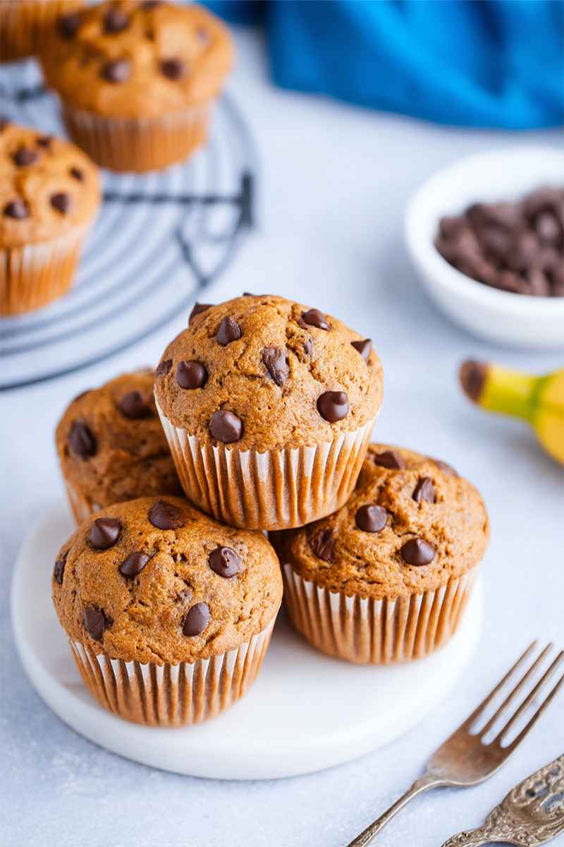 A stack of golden-brown banana chocolate chip muffins on a white plate, surrounded by a bowl of chocolate chips and a banana.