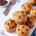 Golden-brown banana chocolate chip muffins on a white plate, with scattered chocolate chips and vintage forks nearby.