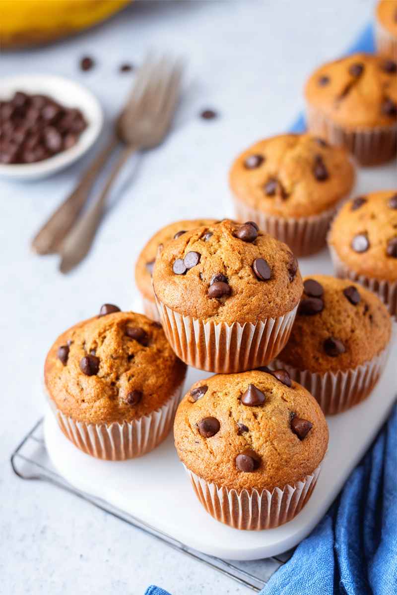 Golden-brown banana chocolate chip muffins on a white plate, with scattered chocolate chips and vintage forks nearby.