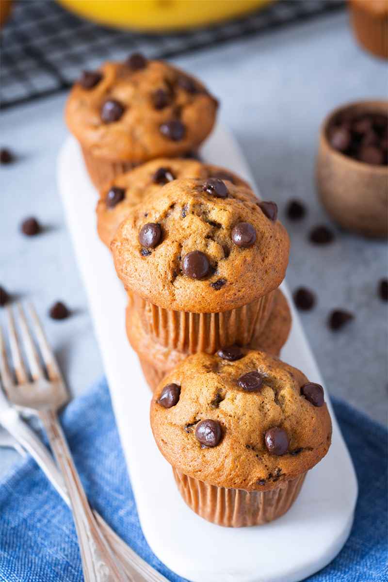 A row of golden-brown banana chocolate chip muffins on a white plate, with scattered chocolate chips and vintage forks nearby.