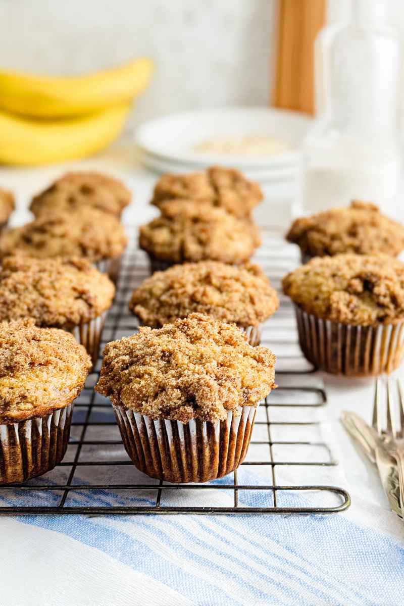 A cooling rack filled with golden-brown banana crumb muffins, set on a blue-striped napkin with bananas and plates in the background.