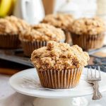 Golden-brown banana crumb muffins with a crunchy topping, served on a white plate with a rustic wooden backdrop.