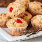 A stack of golden-brown banana pineapple muffins topped with shredded coconut and maraschino cherries, served on a white plate.