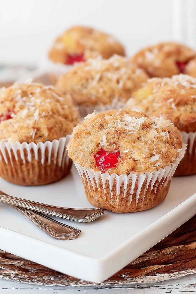 A plate of banana pineapple muffins with coconut shreds and maraschino cherries, placed on a rustic wooden surface.