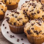 Golden-brown chocolate chip muffins topped with mini chocolate chips, served on a white plate.