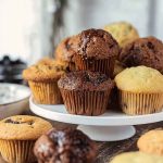 Stacked chocolate muffins with some drizzled in glaze, set on a white cake stand.
