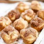 Golden-brown snickerdoodle muffins dusted with cinnamon sugar, served on a white plate with a rustic wooden spoon.