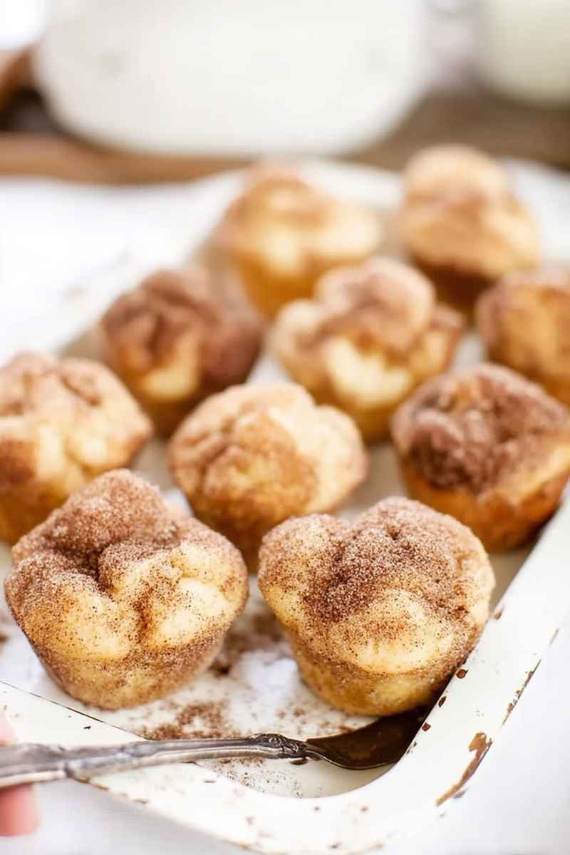 Golden-brown snickerdoodle muffins dusted with cinnamon sugar, served on a white plate with a rustic wooden spoon.