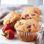 Golden-brown strawberry cream muffins on a white plate, with a whole strawberry and vintage forks nearby.