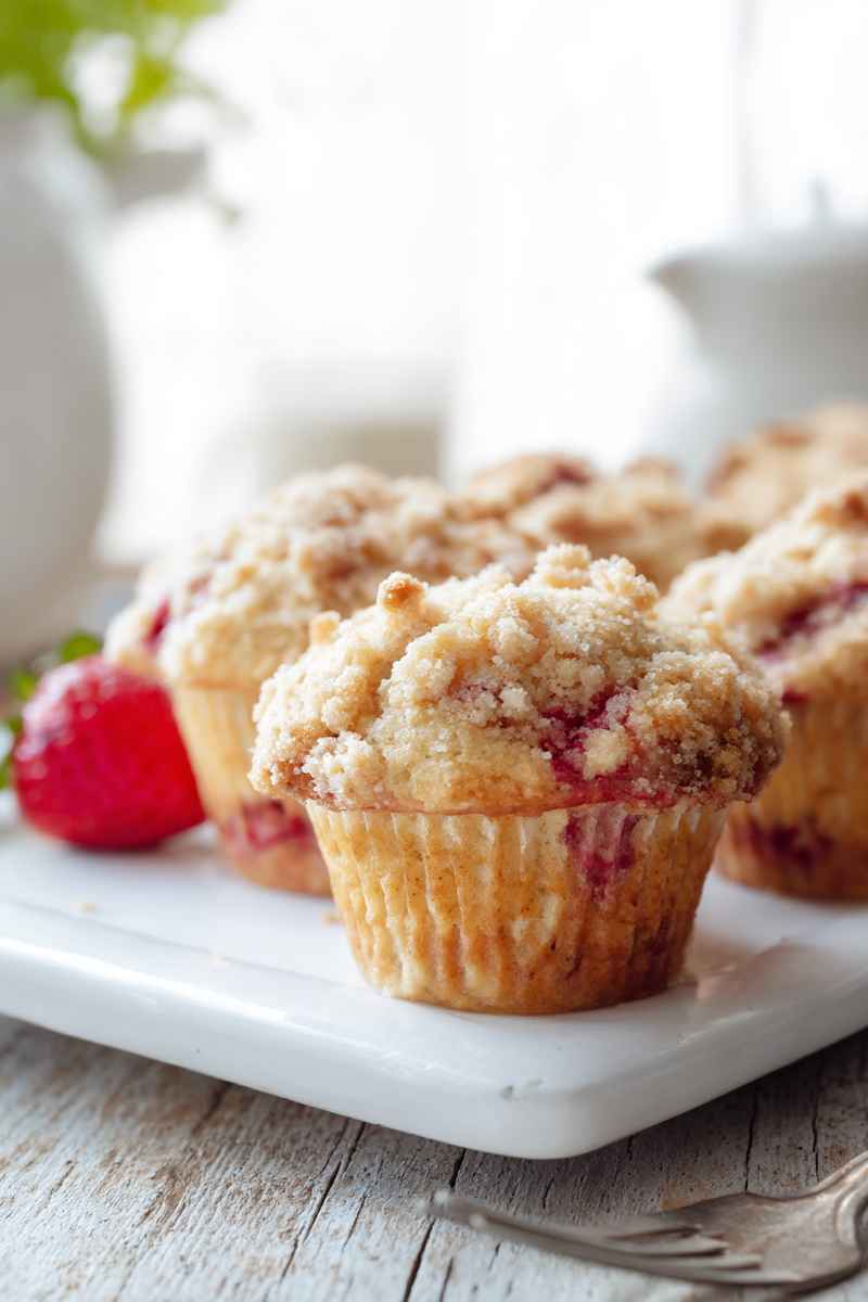 A close-up of a Strawberry Streusel Muffin with a crumbly streusel topping, accompanied by a fresh strawberry.