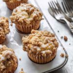 Golden apple muffins with a crumbly streusel topping and a drizzle of creamy glaze, served on a white marble tray with vintage forks.