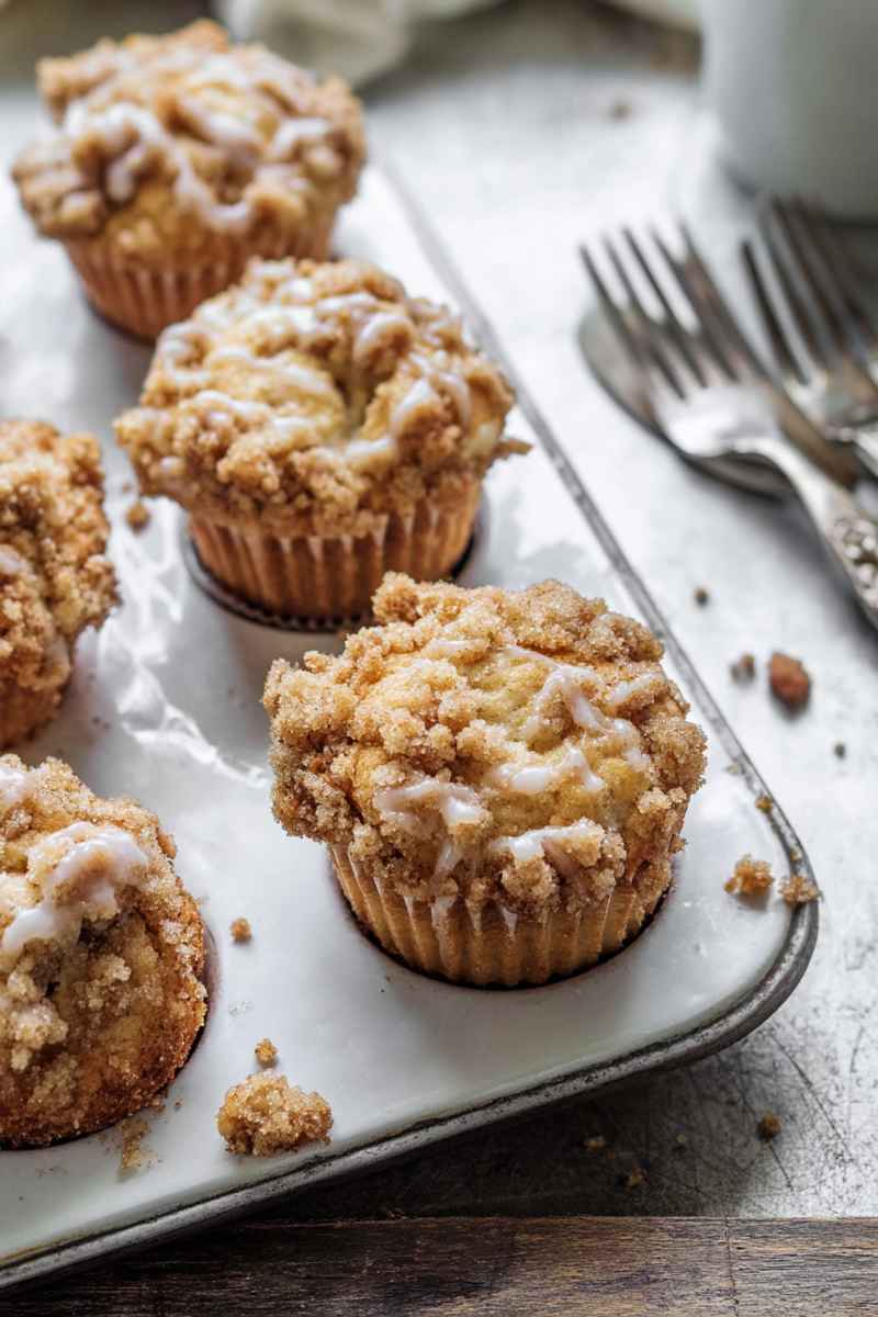 Golden apple muffins with a crumbly streusel topping and a drizzle of creamy glaze, served on a white marble tray with vintage forks.