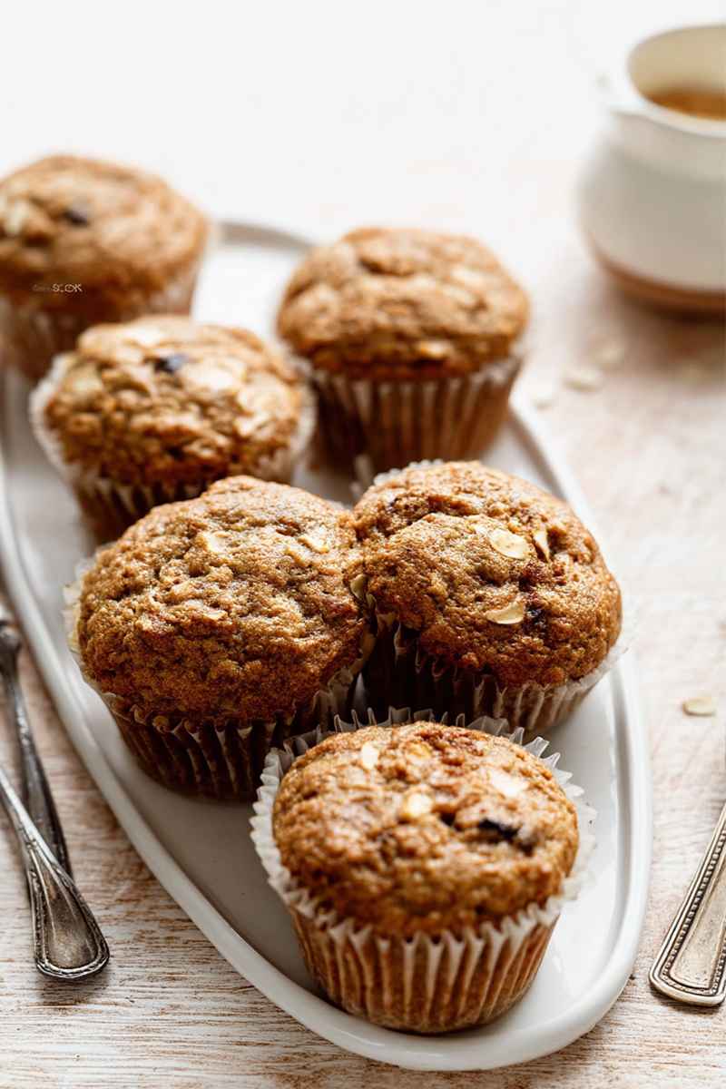 A tray of freshly baked applesauce muffins topped with rolled oats, placed on a rustic table with a cup of coffee in the background.