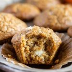 A partially eaten carrot cake muffin with a visible cream cheese filling sits on a silver plate among other whole muffins.