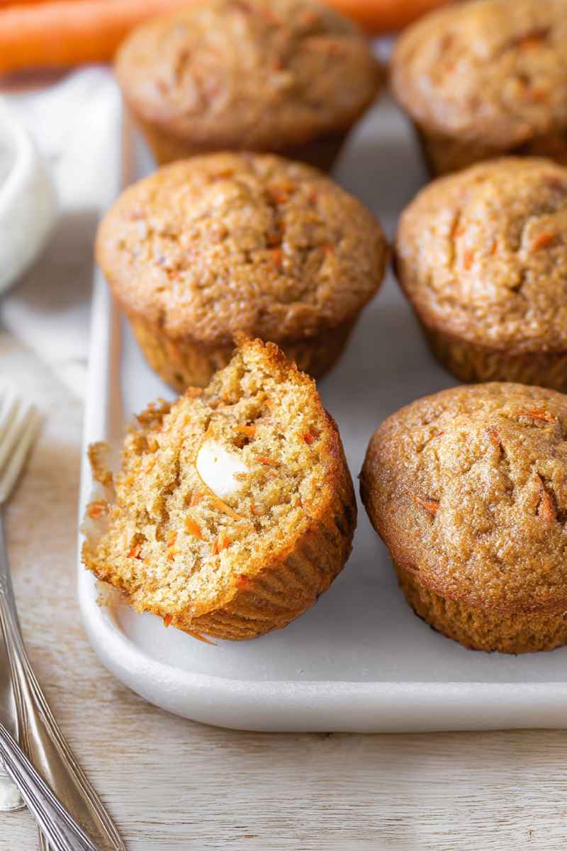 Several carrot cake muffins, one with a bite taken out showing the cream cheese filling, rest on a light-colored surface with forks.