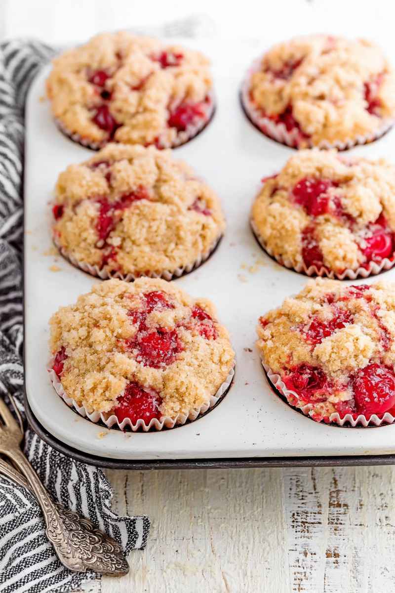 A tray of golden-brown cherry cobbler muffins with visible streusel topping and bright red cherries.
