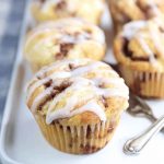 Close-up of glazed cinnamon swirl muffins in paper liners on a white plate with a blue checkered cloth.