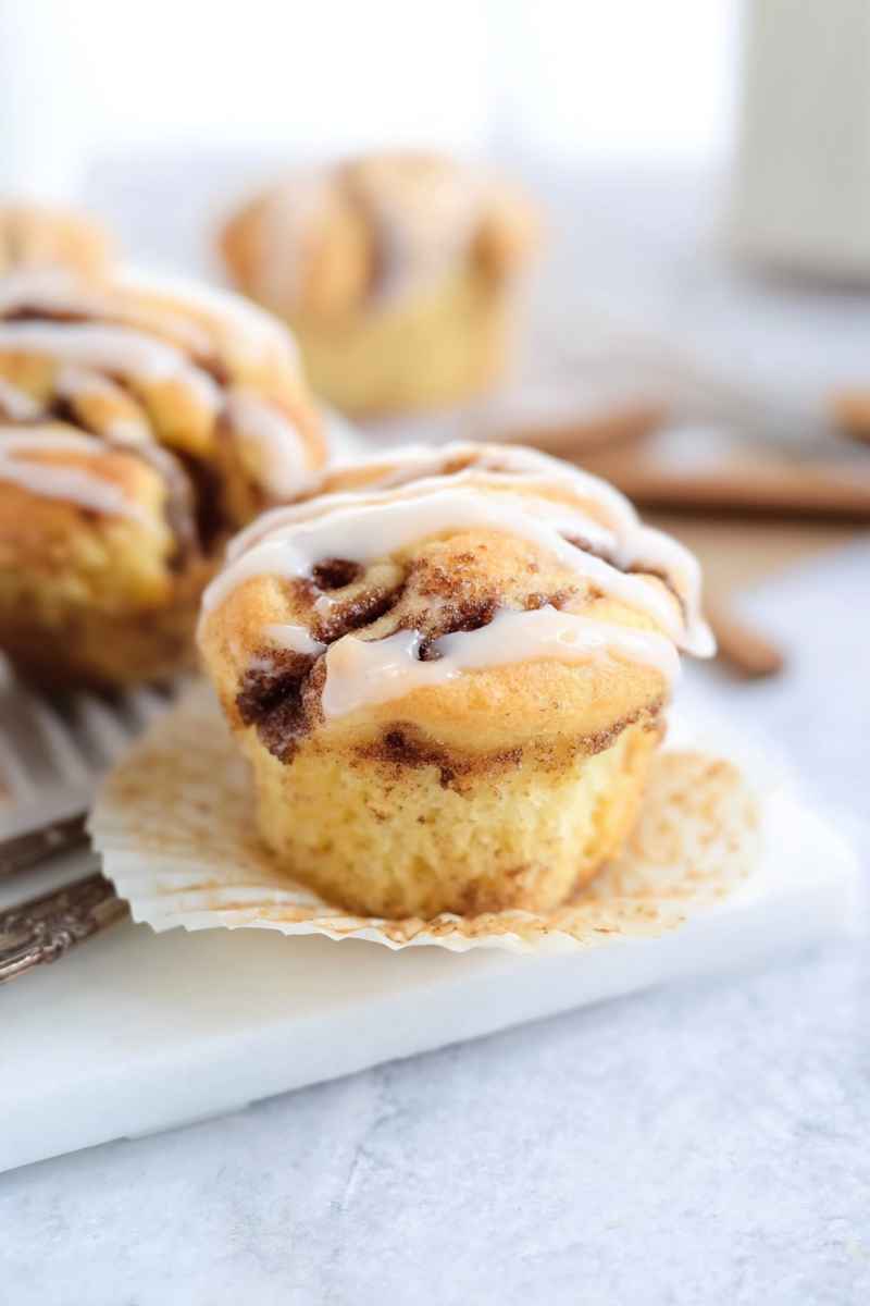 Single glazed cinnamon roll muffin in a white paper liner on a white surface with a fork beside it.