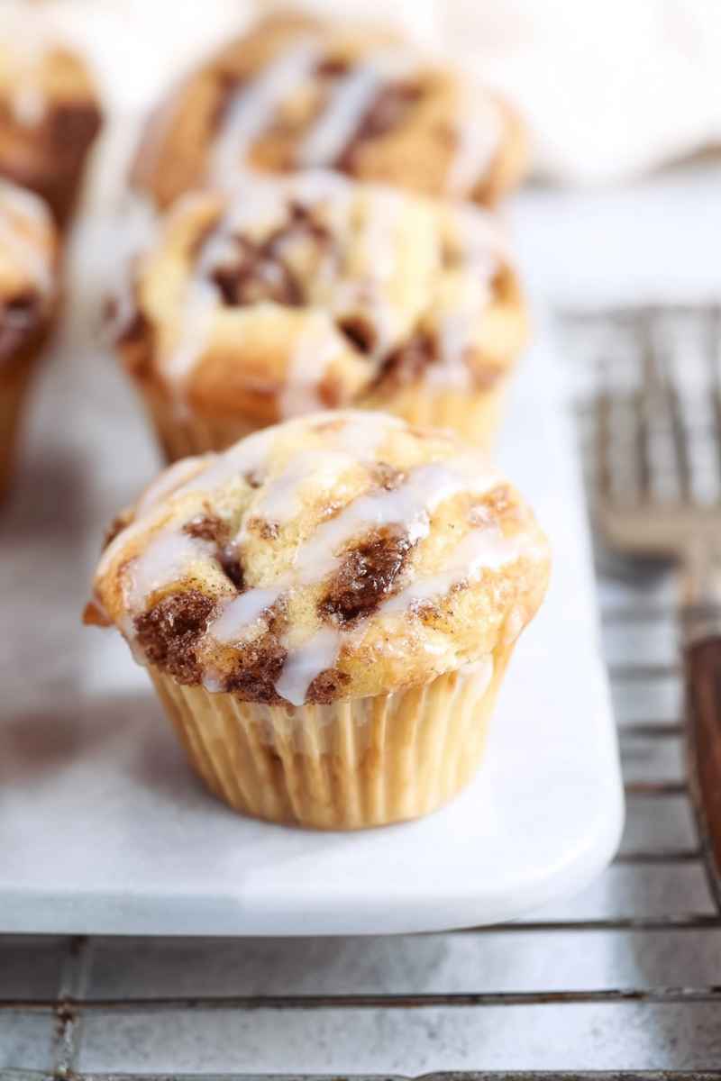 Close-up of a glazed cinnamon streusel muffin with others blurred in the background, on a white surface.