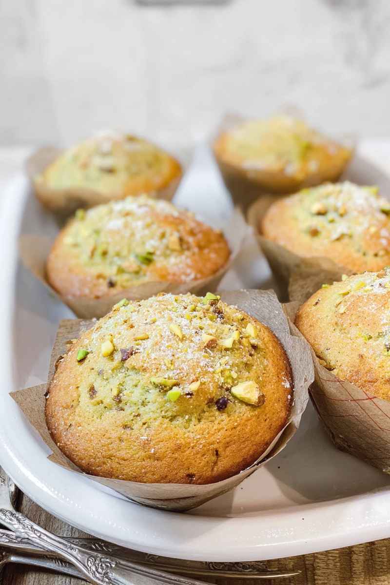 Golden pistachio muffins with a sprinkle of powdered sugar and chopped pistachios, arranged on a white plate.
