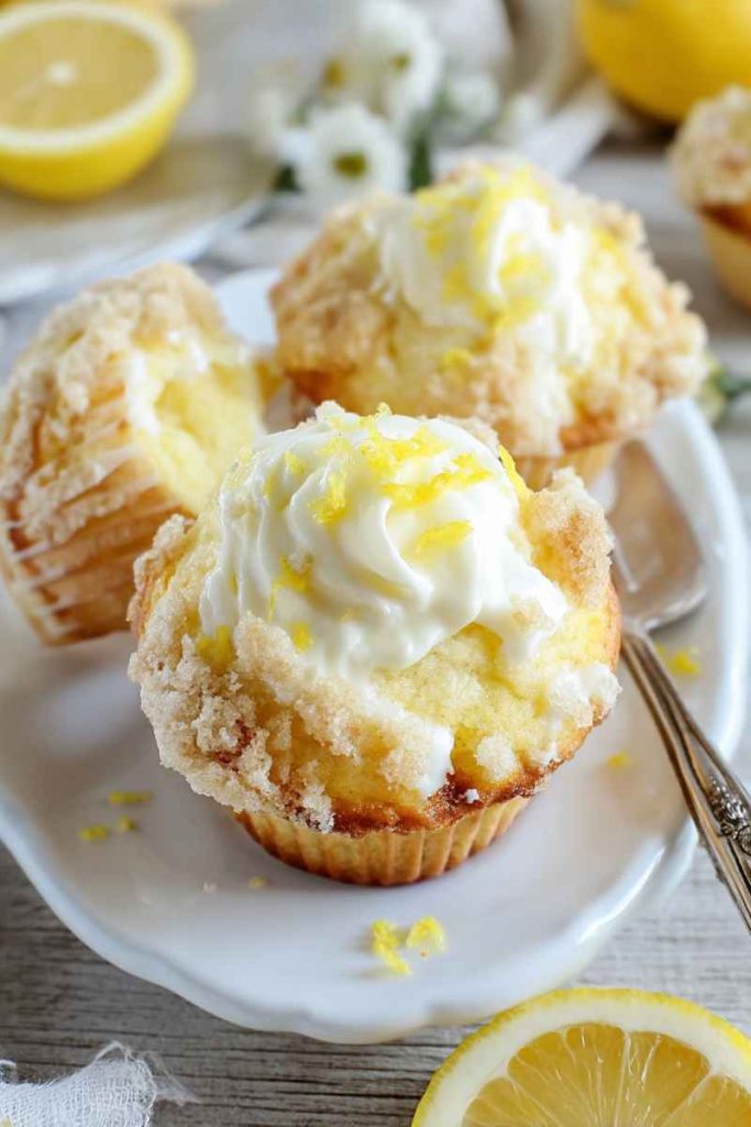 Luscious lemon muffins topped with whipped cream and lemon zest, surrounded by fresh lemons on a white plate.