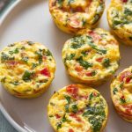 Mini frittata muffins with visible green herbs and red tomato pieces, arranged on a white plate, ready to serve.