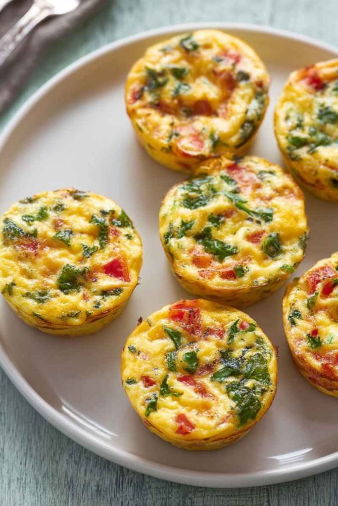 Mini frittata muffins with visible green herbs and red tomato pieces, arranged on a white plate, ready to serve.
