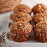 A cluster of carrot muffins with visible nut pieces, set on a rustic wooden table with a woven basket in the background.