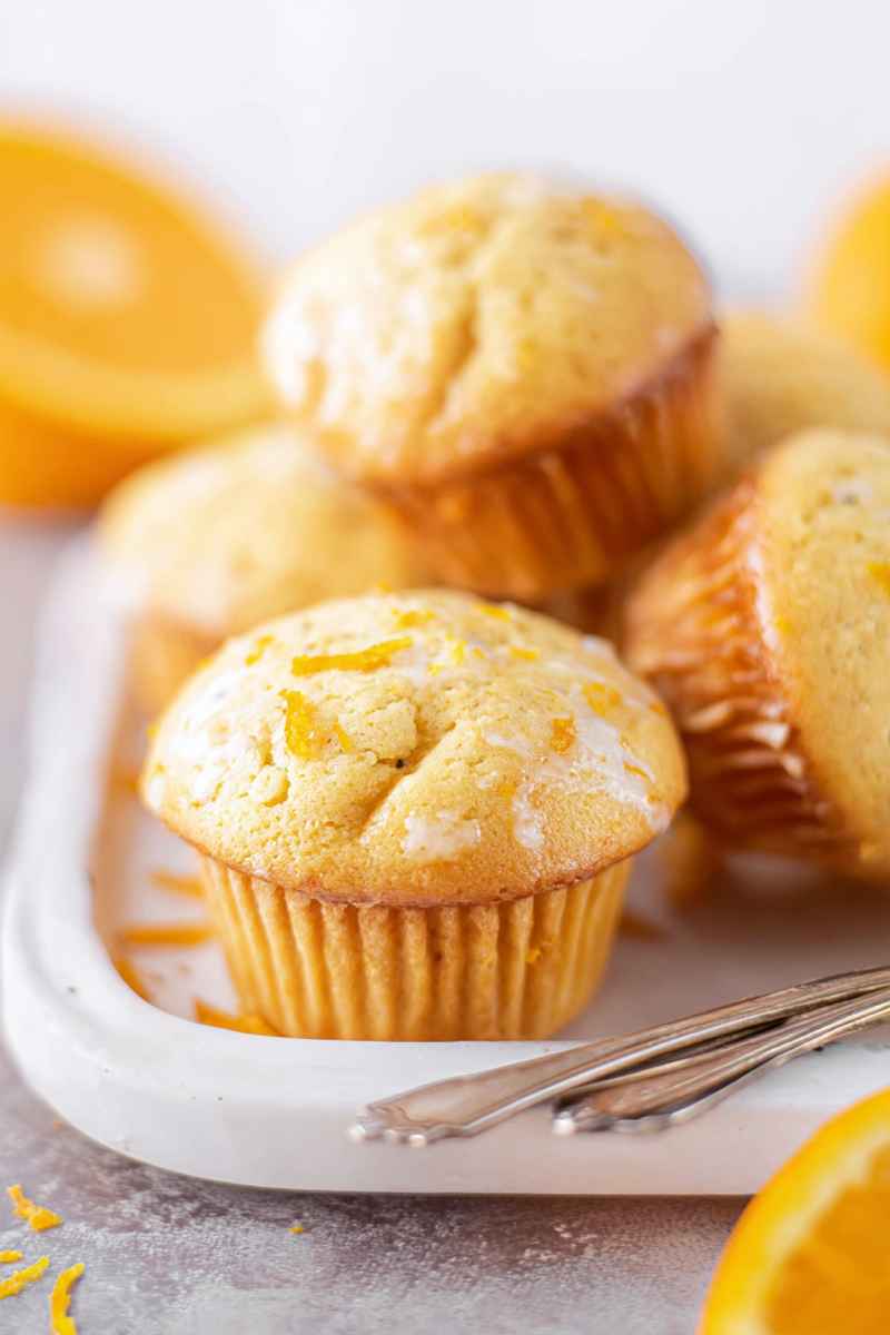 Golden orange muffins arranged on a white plate, drizzled with glaze and surrounded by orange slices.