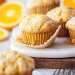 An orange muffin with a shiny glaze and orange zest, displayed on a marble board with forks nearby.