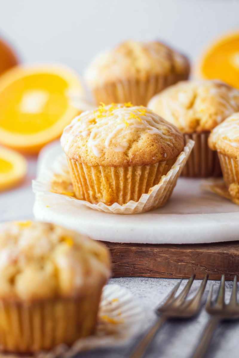 An orange muffin with a shiny glaze and orange zest, displayed on a marble board with forks nearby.