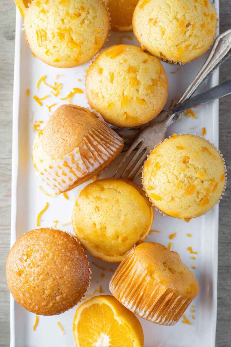 A tray of golden orange muffins sprinkled with zest, accompanied by slices of fresh oranges.