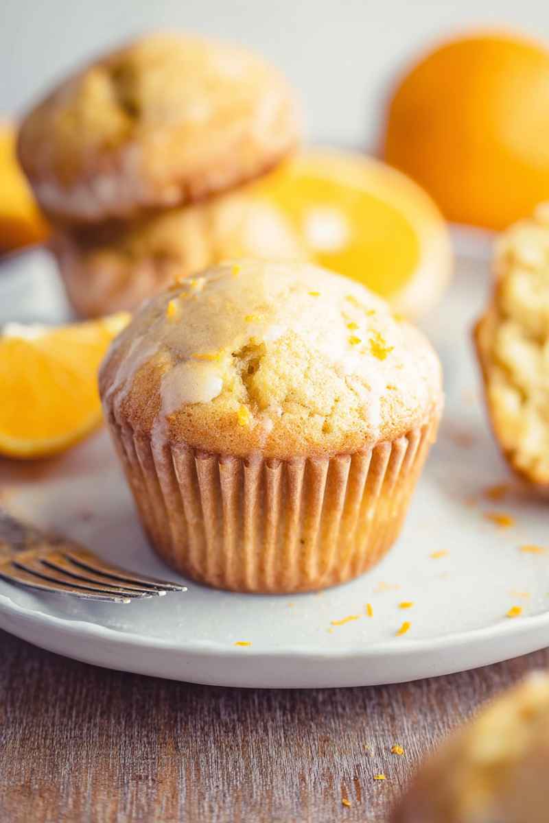 A close-up of an orange muffin with a glossy glaze and orange zest, placed on a white plate.