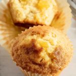 Two golden pineapple muffins in paper liners, placed on a light wooden table, ready to be enjoyed.