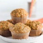 Four zucchini carrot oatmeal muffins in white paper liners, arranged on a white cake stand, with a blurred carrot in the background.