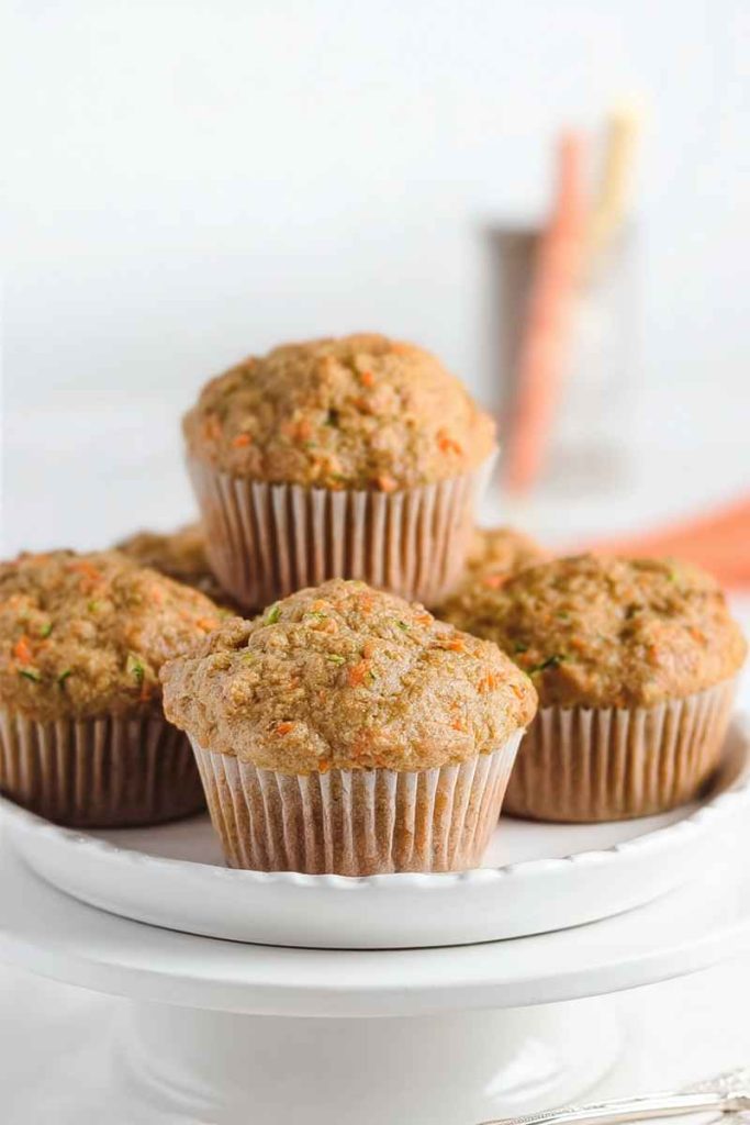 Four zucchini carrot oatmeal muffins in white paper liners, arranged on a white cake stand, with a blurred carrot in the background.