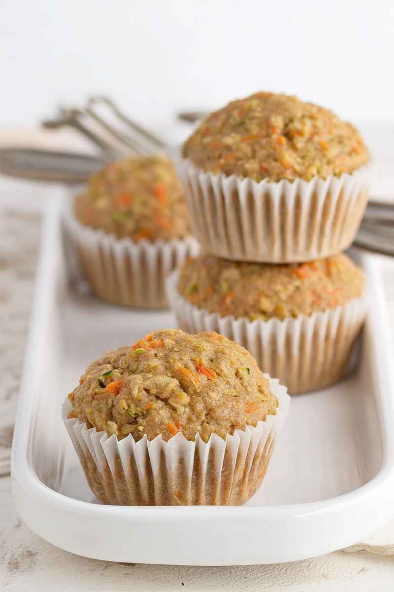 Three zucchini carrot oatmeal muffins in white paper liners, placed on a white tray, with a fork in the background.