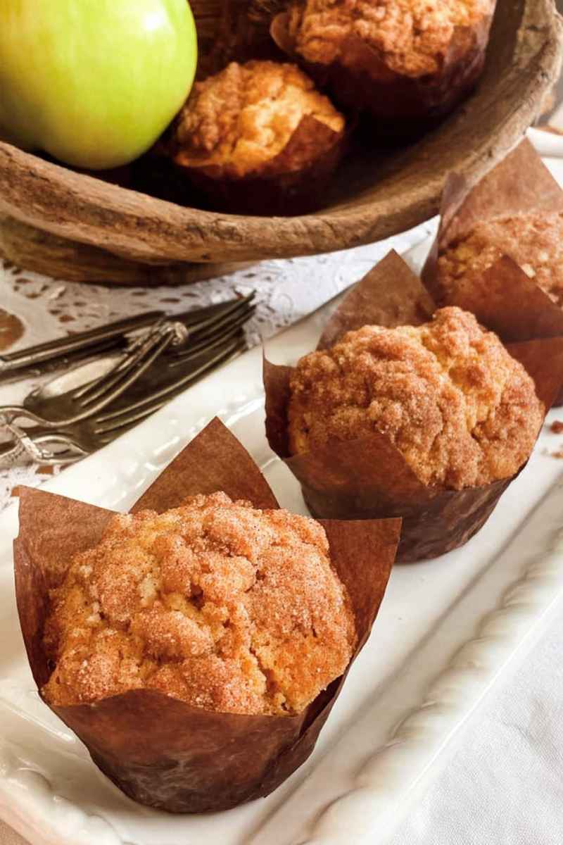 Golden-brown apple cinnamon muffins in brown paper liners on a white plate, with fresh apples and rustic decor in the background.
