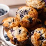 A close-up of golden, fluffy blueberry muffins with visible blueberries peeking through, arranged on a white plate with fresh blueberries scattered around.