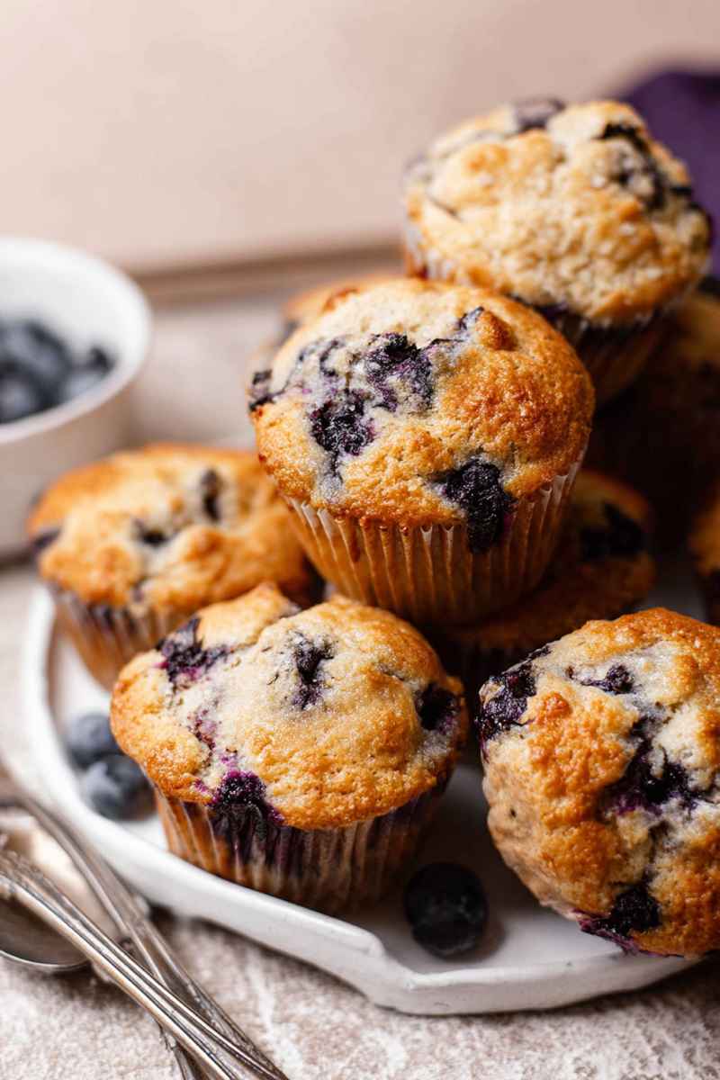 A close-up of golden, fluffy blueberry muffins with visible blueberries peeking through, arranged on a white plate with fresh blueberries scattered around.
