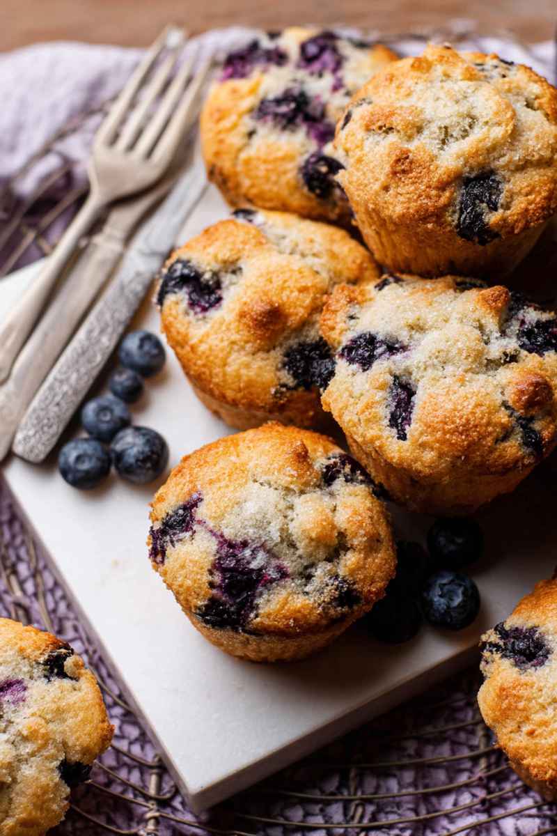 A stack of golden-brown blueberry muffins with visible juicy blueberries and a sprinkling of lemon sugar on top, placed on a white tray with fresh blueberries scattered around.