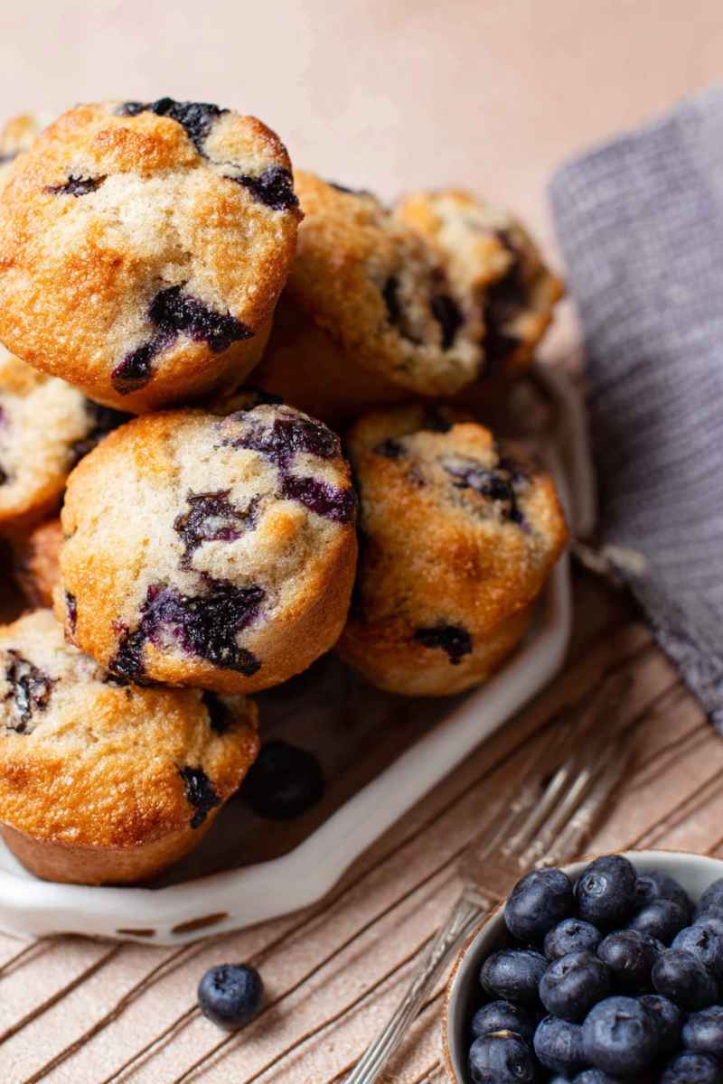 A plate of blueberry muffins with a crisp, sugary topping, surrounded by fresh blueberries and a cozy kitchen setting.