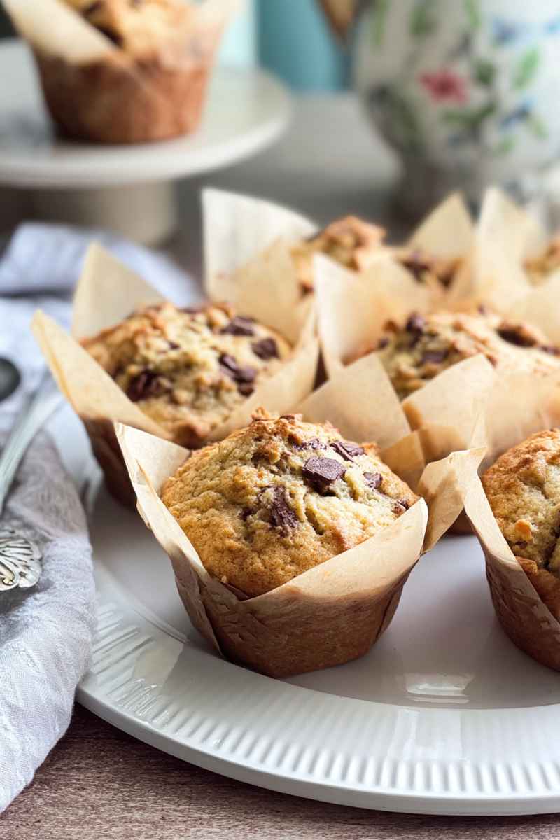 Delicious banana muffins topped with chocolate chunks, arranged on a white plate with brown paper liners.