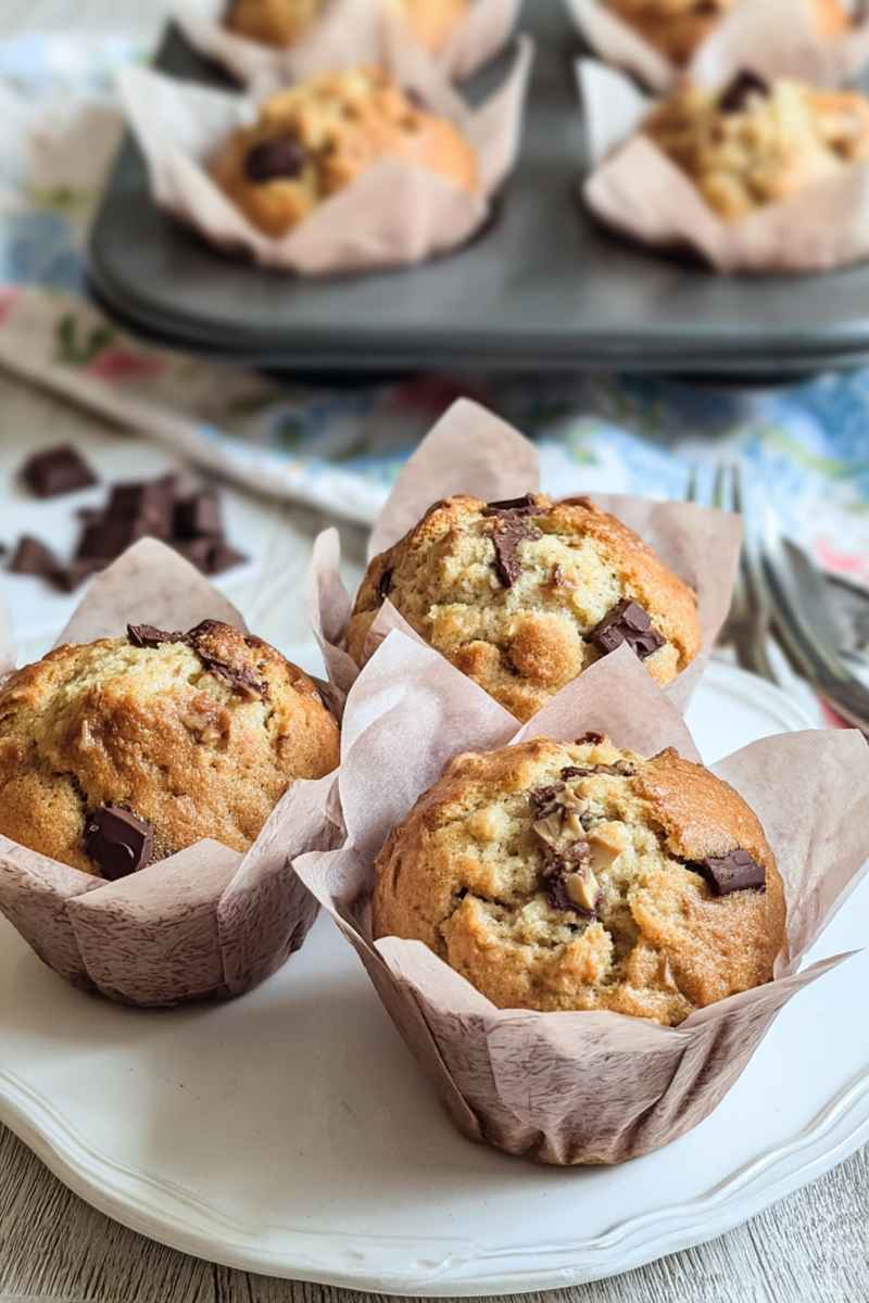 A close-up of banana muffins with visible chocolate chunks, placed on a plate with floral-patterned paper liners.