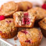 A plate of golden-brown banana raspberry muffins with one halved to reveal a juicy raspberry filling.